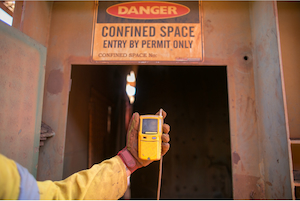 Worker holding a portable gas monitor outside a permit-required confined space entrance.