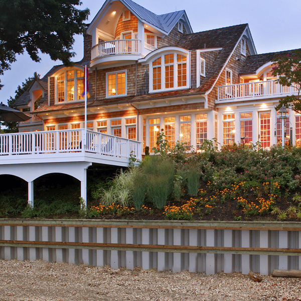 Residential backyard with green lawn, trees, patio seating, and a seawall next to calm water.