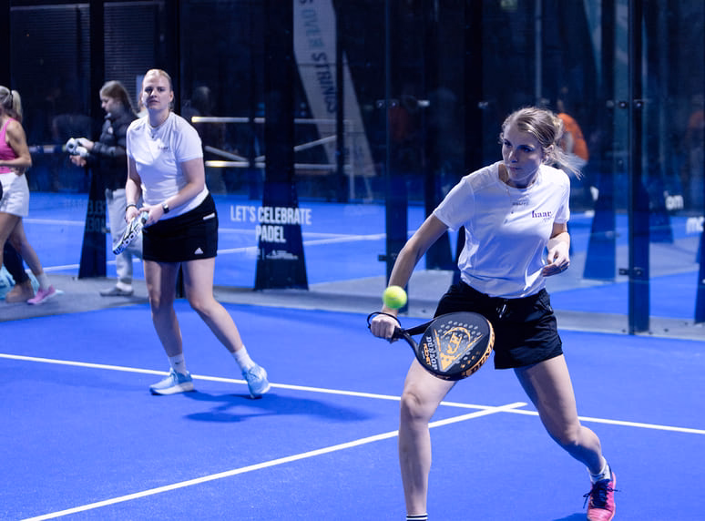 Vrouwelijke padelspelers in actie op een indoorbaan tijdens een evenement met ondersteuning van Haarspullen.nl.