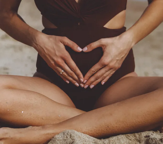 A woman sitting on the beach crosslegged, with her hands held over her abdomen in the shape of a heart for a story on pelvic self-exam.