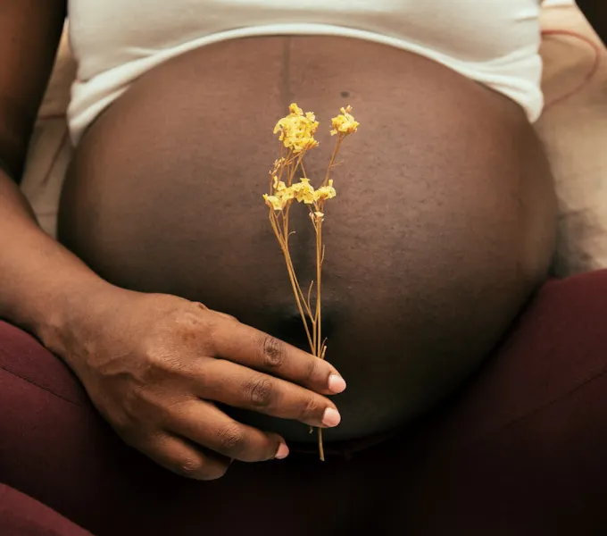 A black pregnant woman holding a small yellow flower in front of her belly.