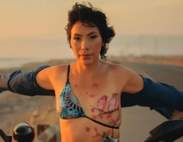 A breast cancer survior sitting confidently astride a motorcycle with a tattoo of birds over her surgery scar.