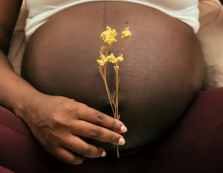 A black pregnant woman holding a small yellow flower in front of her belly.