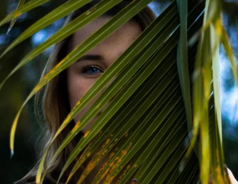 A woman standing behind a large plant so that only her bright blue eye is visble.