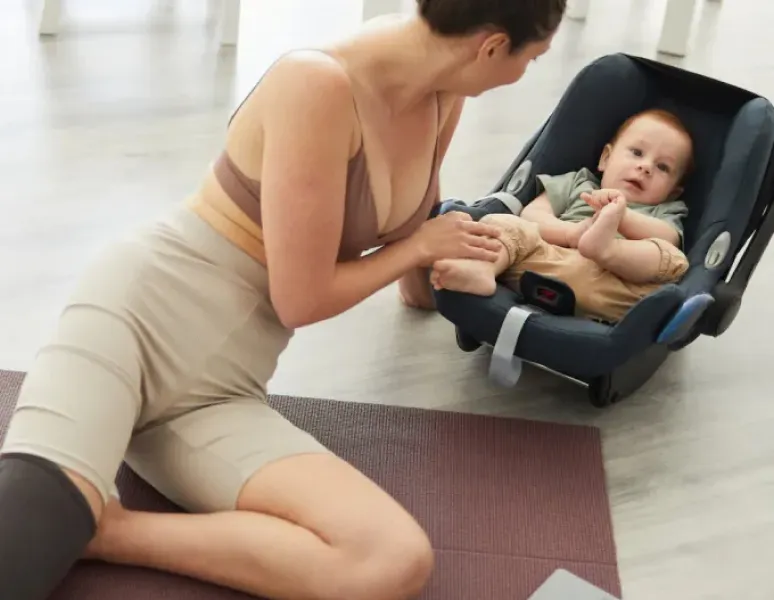 A woman on a yoga mat leaning over to say hi to her baby in a car seat. a laptop is open for her virtual postpartum physical therapy visit.