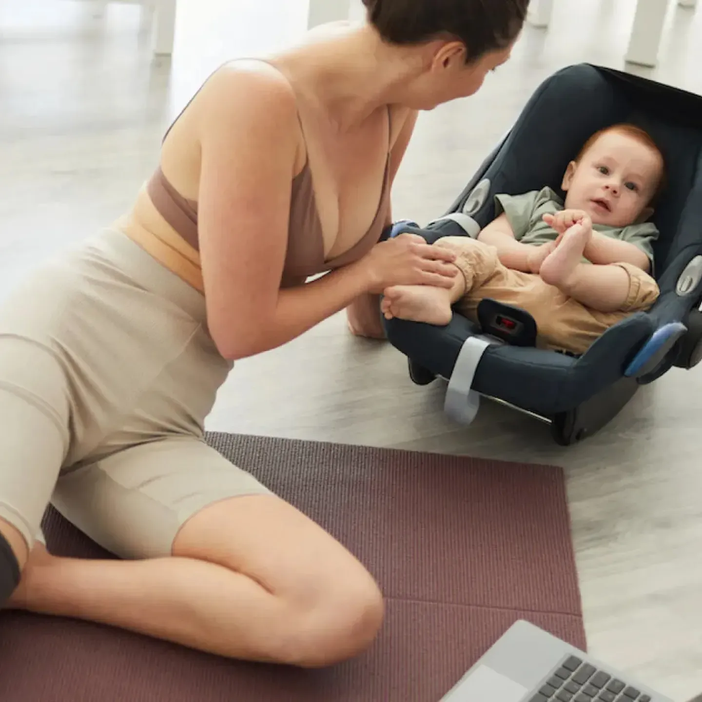 A woman on a yoga mat leaning over to say hi to her baby in a car seat. a laptop is open for her virtual postpartum physical therapy visit.