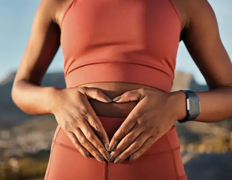 A woman wearing a rust colored t-shirt knotted at the waist, holding a pink exercise ball above her head to strengthen her deep core with exercise.