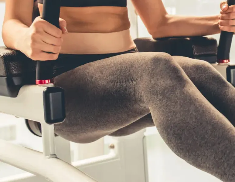 A woman in gray leggings and a pink sports bra having a coregasm while using an ab exercise machine at the gym.