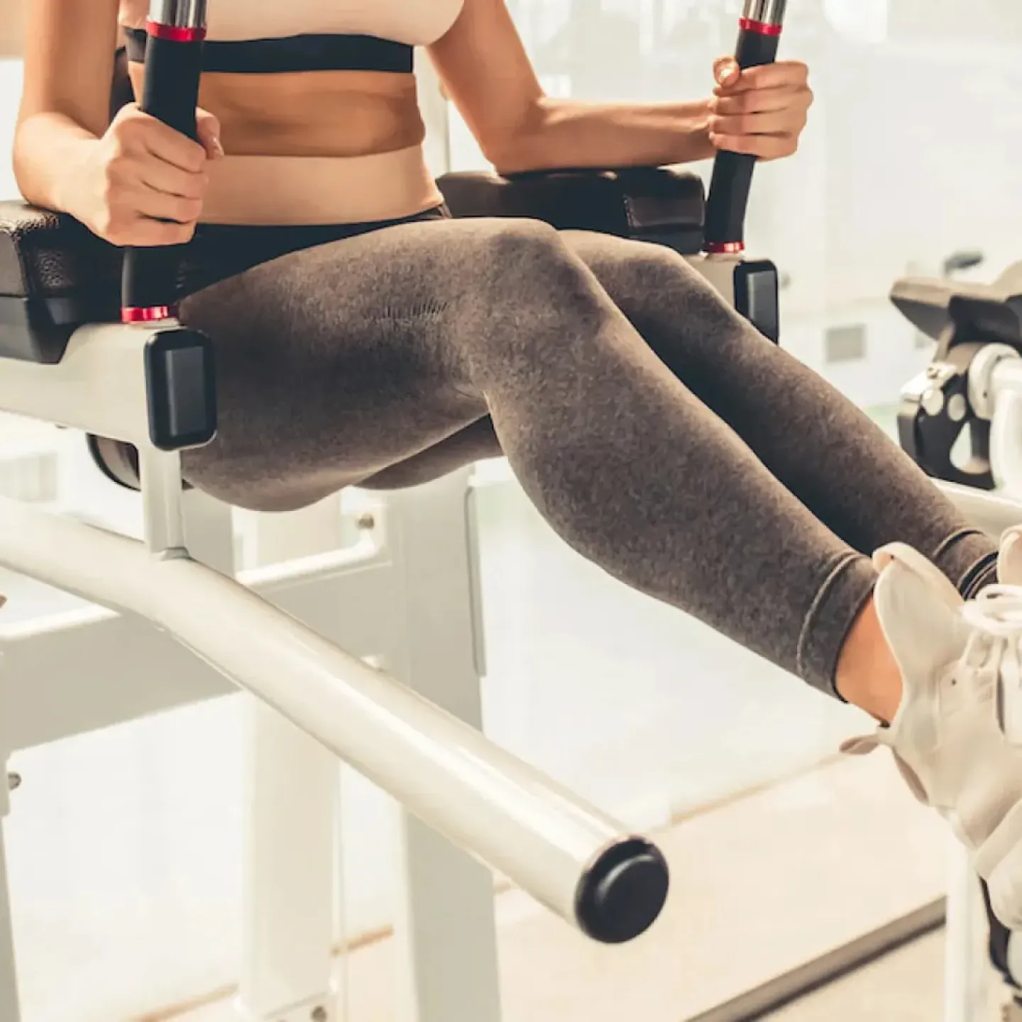 A woman in gray leggings and a pink sports bra having a coregasm while using an ab exercise machine at the gym.
