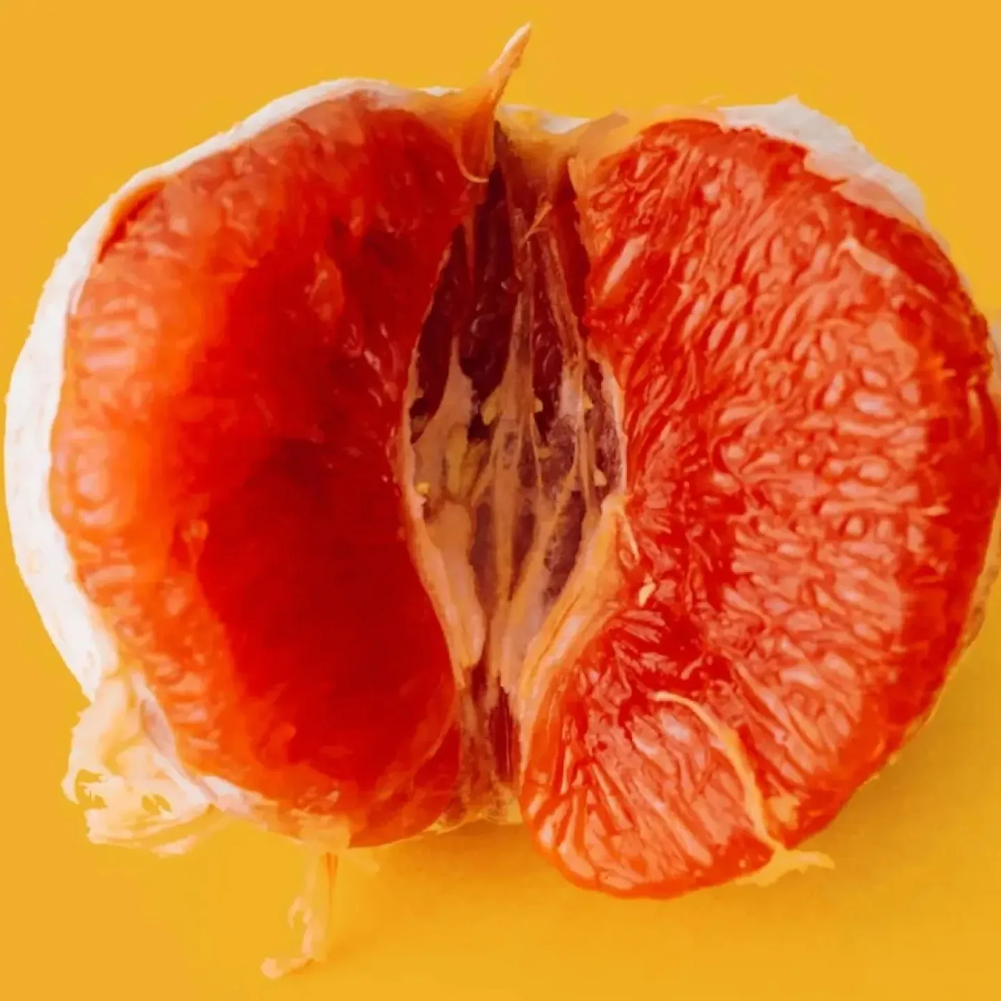 A close-up photo of a peeled, halved blood orange on a mustard yellow background to symbolize high genital esteem and big vulva energy.