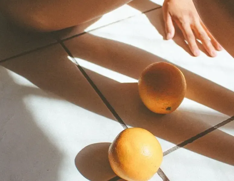 A woman sitting in the sun next to two oranges with artful shadows falling on the floor for story about hysterectomy recovery time.