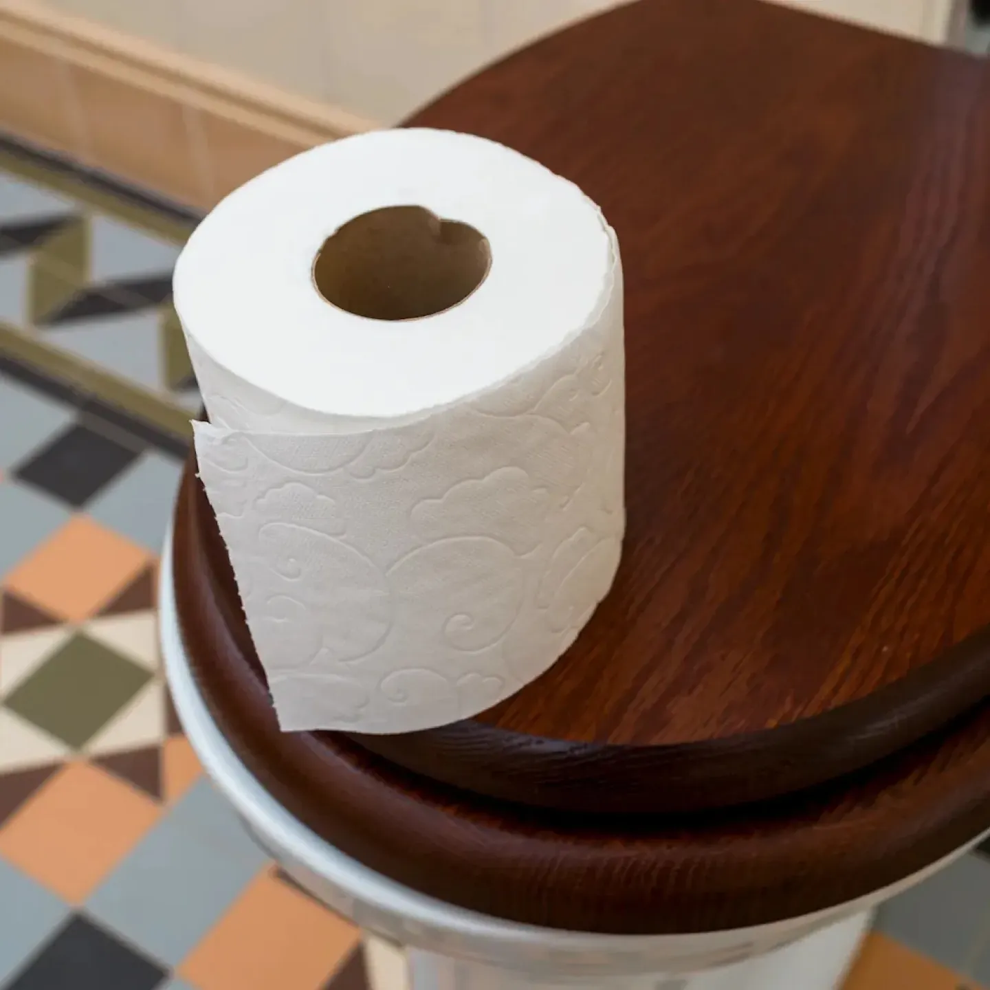 A roll of toilet paper on a wooden toilet seat in a bathroom with colorful tiles.