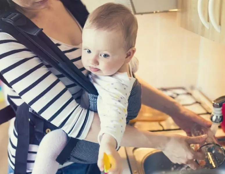 A new mom with post c-section back pain washing dishes while holding her baby in a carrier.