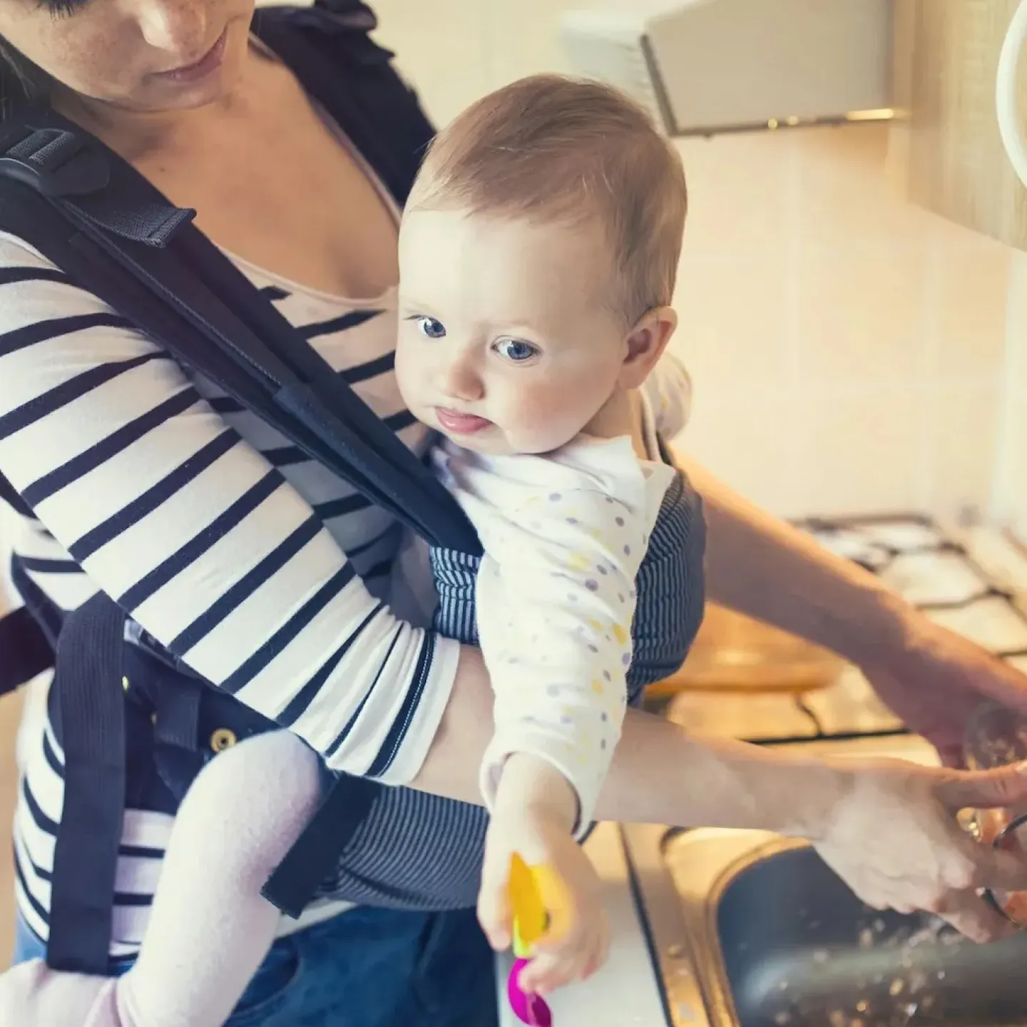 A new mom with post c-section back pain washing dishes while holding her baby in a carrier.