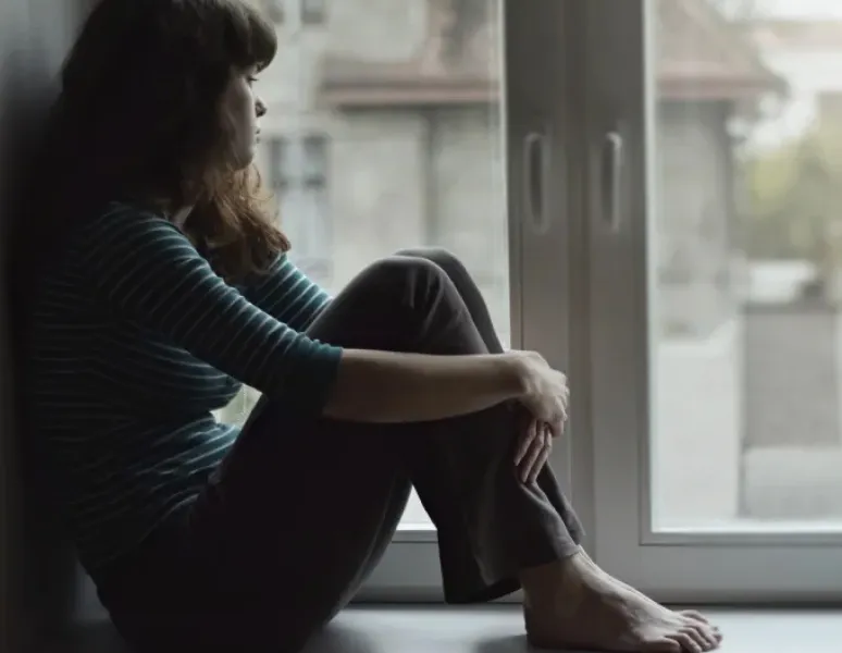 Sad looking woman sitting on a windowsill looking out the window.