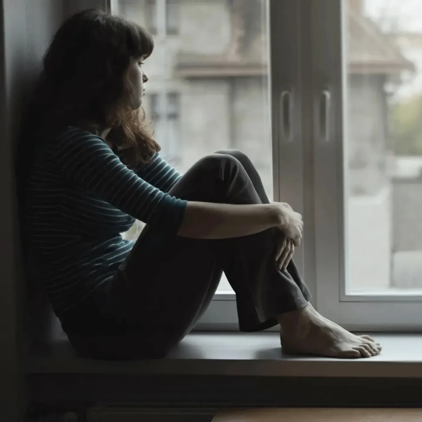 Sad looking woman sitting on a windowsill looking out the window.