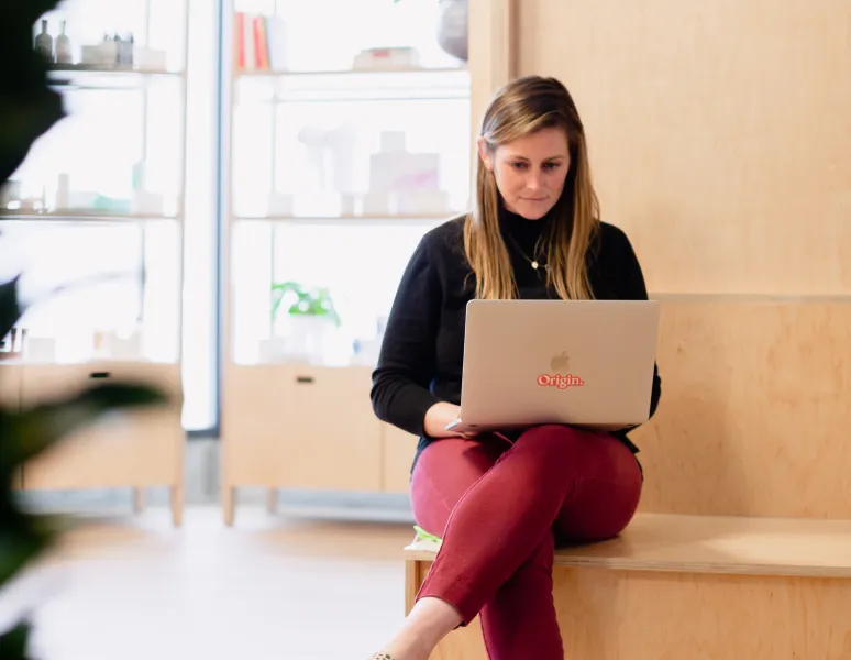 A physical therapist sits on a bench working on a laptop in a modern, light-filled Origin clinic.