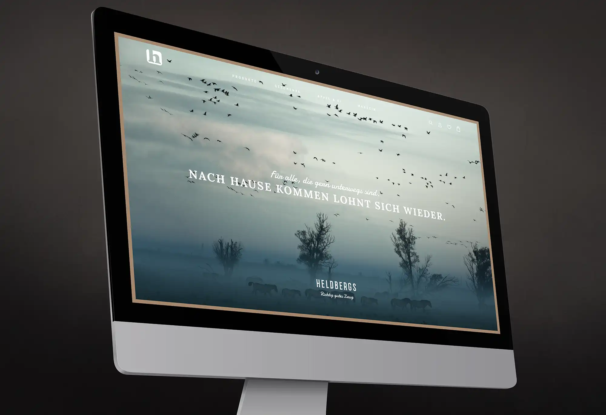 Auf einem großen Bildschirm sehen wir die Startseite des HELDBERGS Online-Shop mit einem nebelverhangenen Bild von Wildpferden und Zugvögeln. Darauf der Text: Für alle, die gern unterwegs sind. Nachhause kommen lohnt sich wieder..