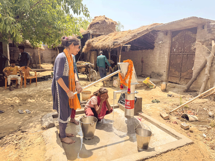 Une jeune femme pompe de l'eau avec une pompe à main, à l'arrière-plan on voit des huttes au toit de chaume.