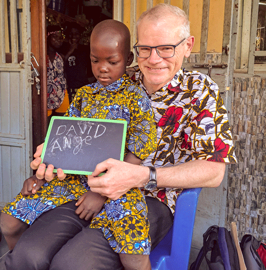 David hat ein Kind auf seinem Schoss, zusammen halten sie eine Schiefertafel mit ihren Namen drauf.
