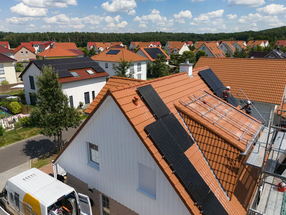 Ein Hausdach mit hellbraunen Ziegeln aus der Vogelperspektive, während eine Solaranlage installiert wird. Man sieht das Montagegerüst, die Montageschienen, zwei Handwerker auf dem Dach und einige fertig montierte Solarmodule.