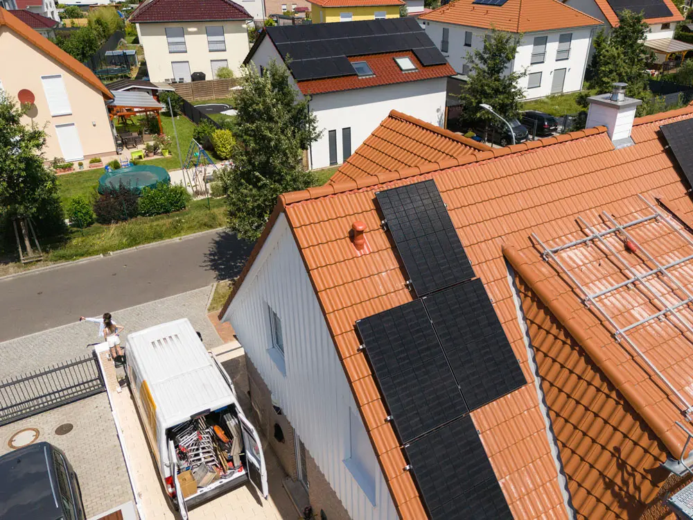 Ein Hausdach mit hellbraunen Ziegeln aus der Vogelperspektive, während eine Solaranlage installiert wird. Man sieht das Montagegerüst, die Montageschienen, zwei Handwerker auf dem Dach und einige fertig montierte Solarmodule.