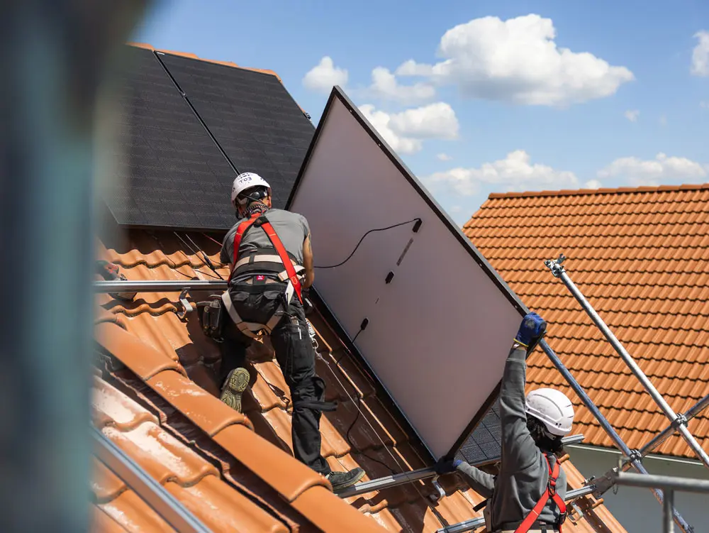 Zwei Enpal Monteure bei der Montage einer PV-Anlage auf einem Hausdach mit hellbraunen Dachziegeln. Man sieht zwei fertig montierte Solarmodule und ein Solarmodul von der Rückseite, während es hochgetragen wird.