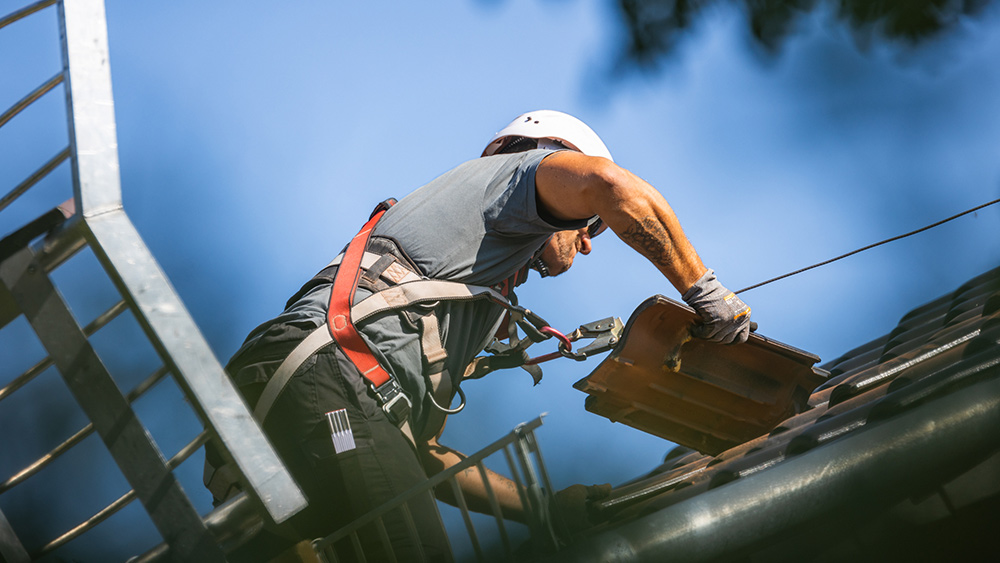 Ein Solarmonteur bereitet die Dachziegel auf einem Hausdach für die Installation einer PV-Anlage vor. An der Seite des Bildes sieht man einen Fallschutz für die Monteure.