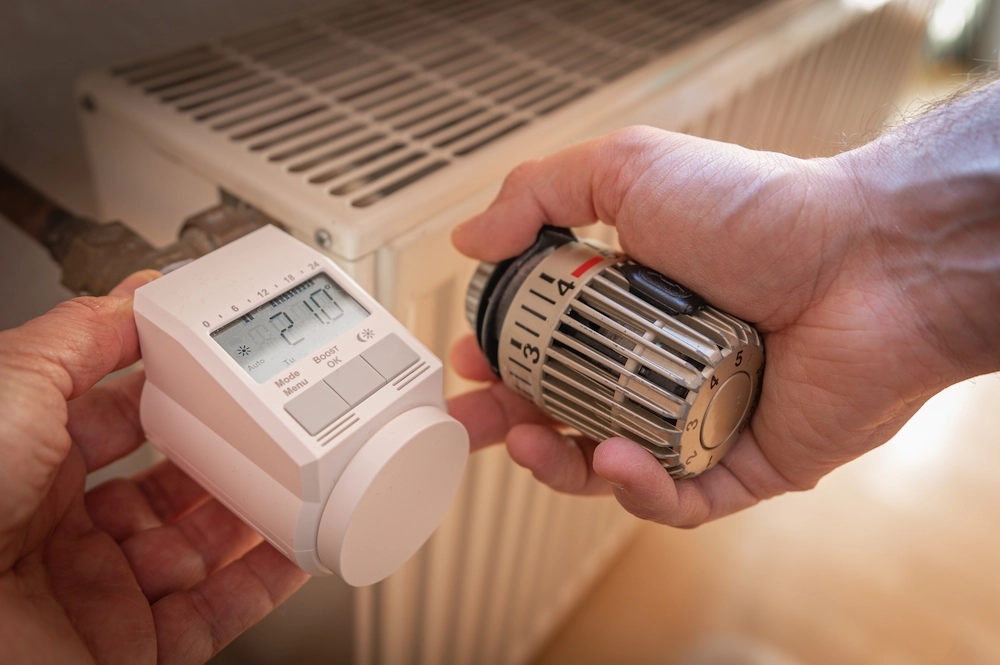 Hände halten ein analoges Thermostat und ein Smart Thermostat vor einer Heizung