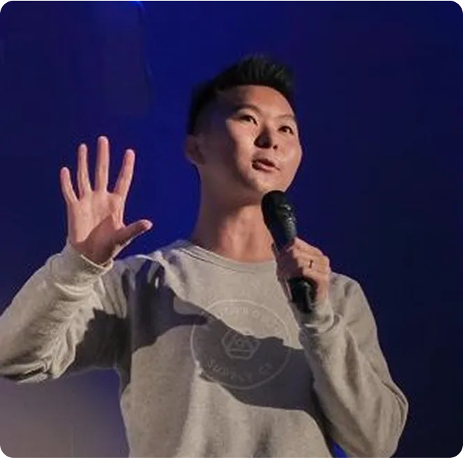 A person wearing a gray sweatshirt holds a microphone and gestures with their right hand, speaking against a blue background.