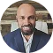 Bald man with beard wearing a navy blazer and white shirt, smiling indoors with blurred background.