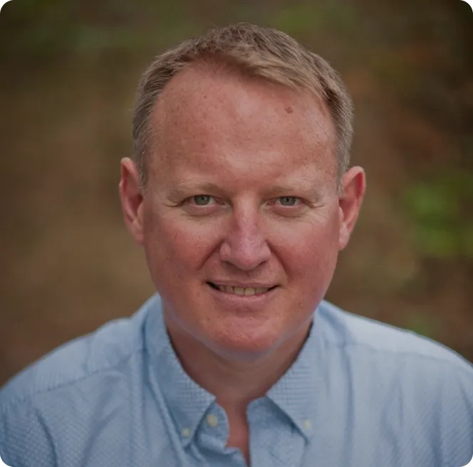 Middle-aged man with short light brown hair wearing a light blue button-up shirt, smiling.