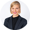 Blonde woman in navy blue blazer looking over her shoulder against a light gray background.
