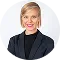 Blonde woman in navy blue blazer looking over her shoulder against a light gray background.