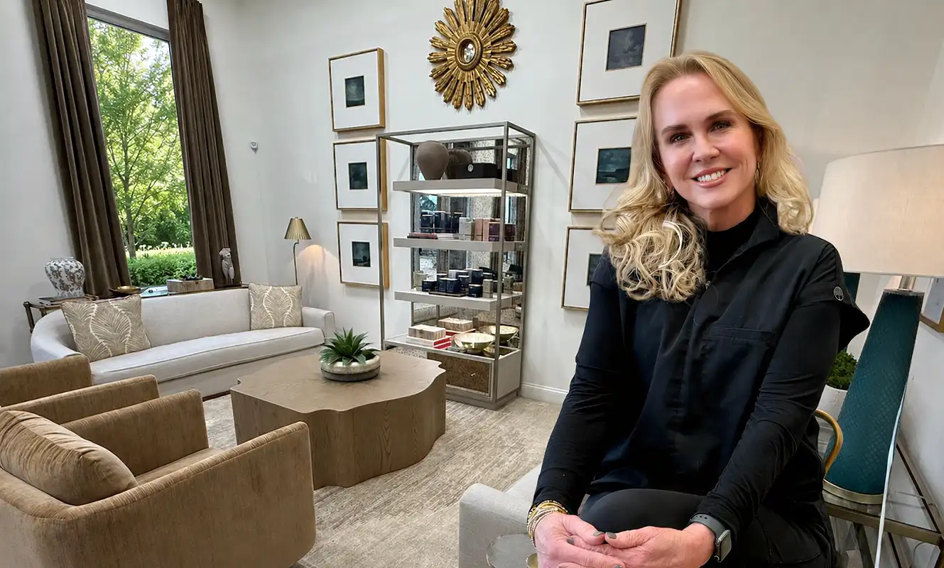 Smiling woman with blonde hair sitting in a stylish living room with beige and white furniture.