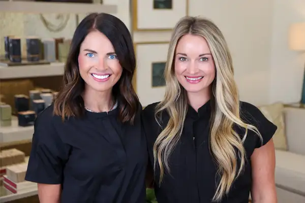 Two smiling women in black medical scrubs standing in a clinic room with a beige chair.