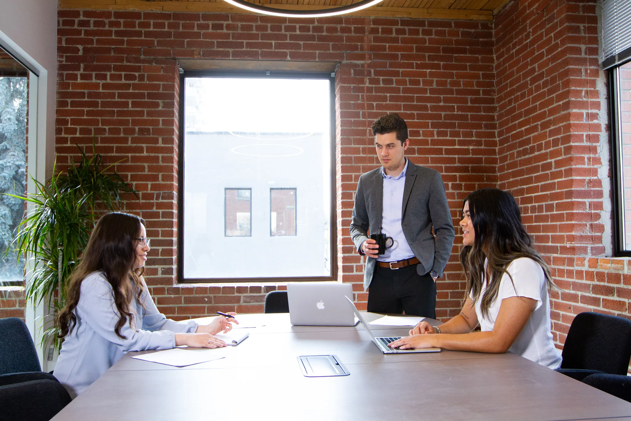 Three professionals having a meeting in a modern office with exposed brick walls, one man standing with a coffee mug, two women sitting with laptops and papers.