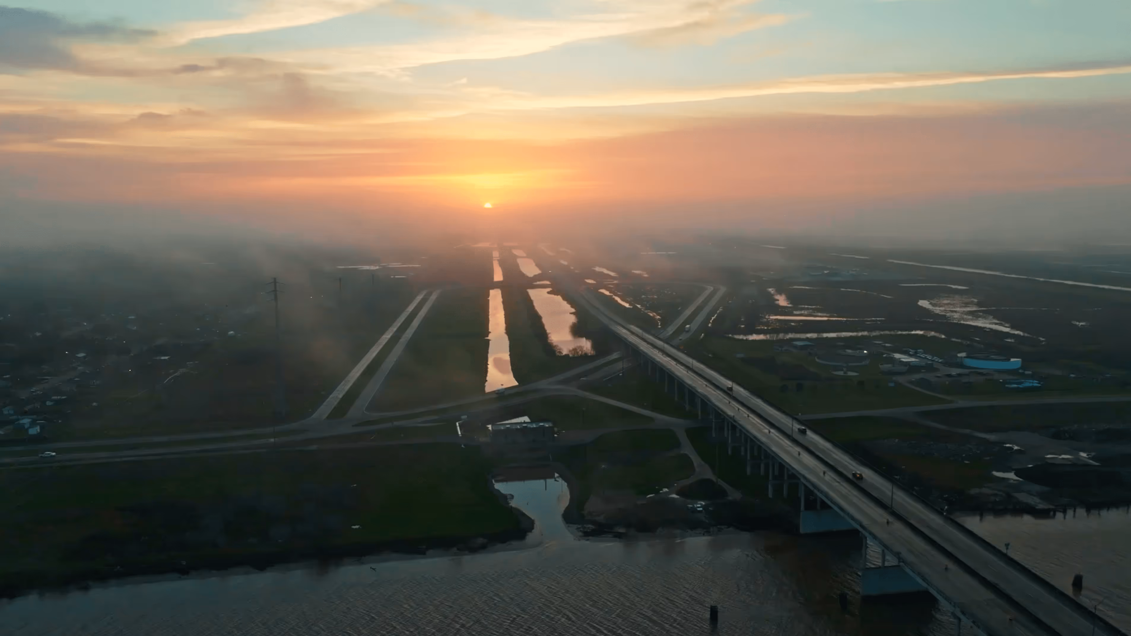 Sunset over a river with a long bridge and surrounding wetlands under a cloudy sky.