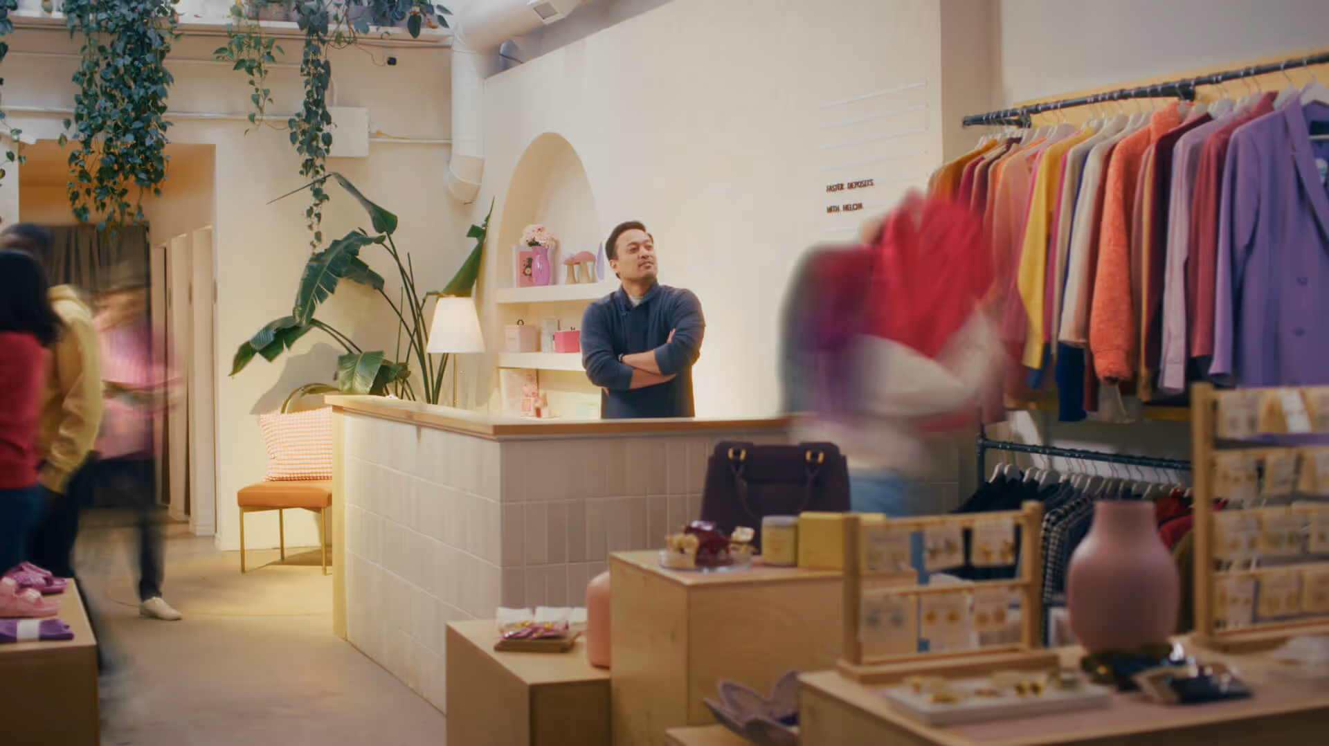 Man in a dark sweater stands behind a shop counter with arms crossed while customers browse colorful clothing on racks.
