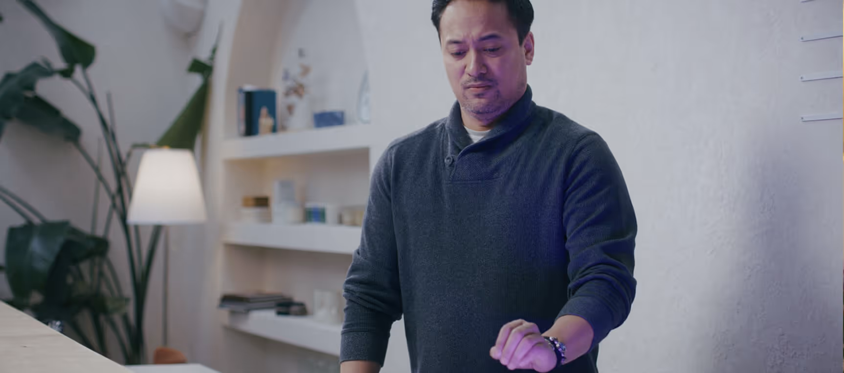 Man in a dark sweater looking at his wristwatch in a modern room with shelves and a floor lamp.