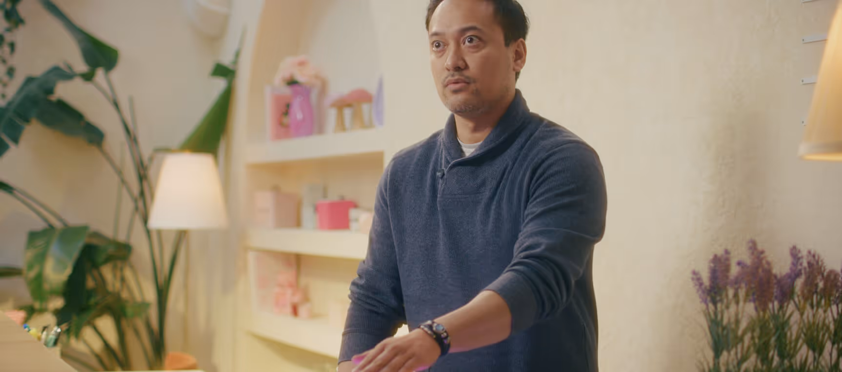 Man in a blue sweater standing indoors with plants, shelves, and lamps in the background.