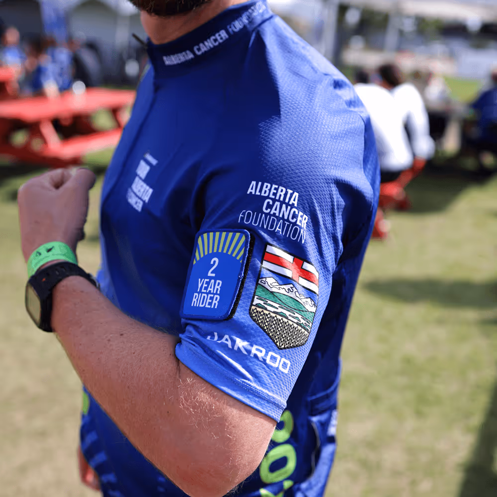 Person wearing a blue cycling jersey with Alberta Cancer Foundation logo and a '2 Year Rider' patch on the sleeve.