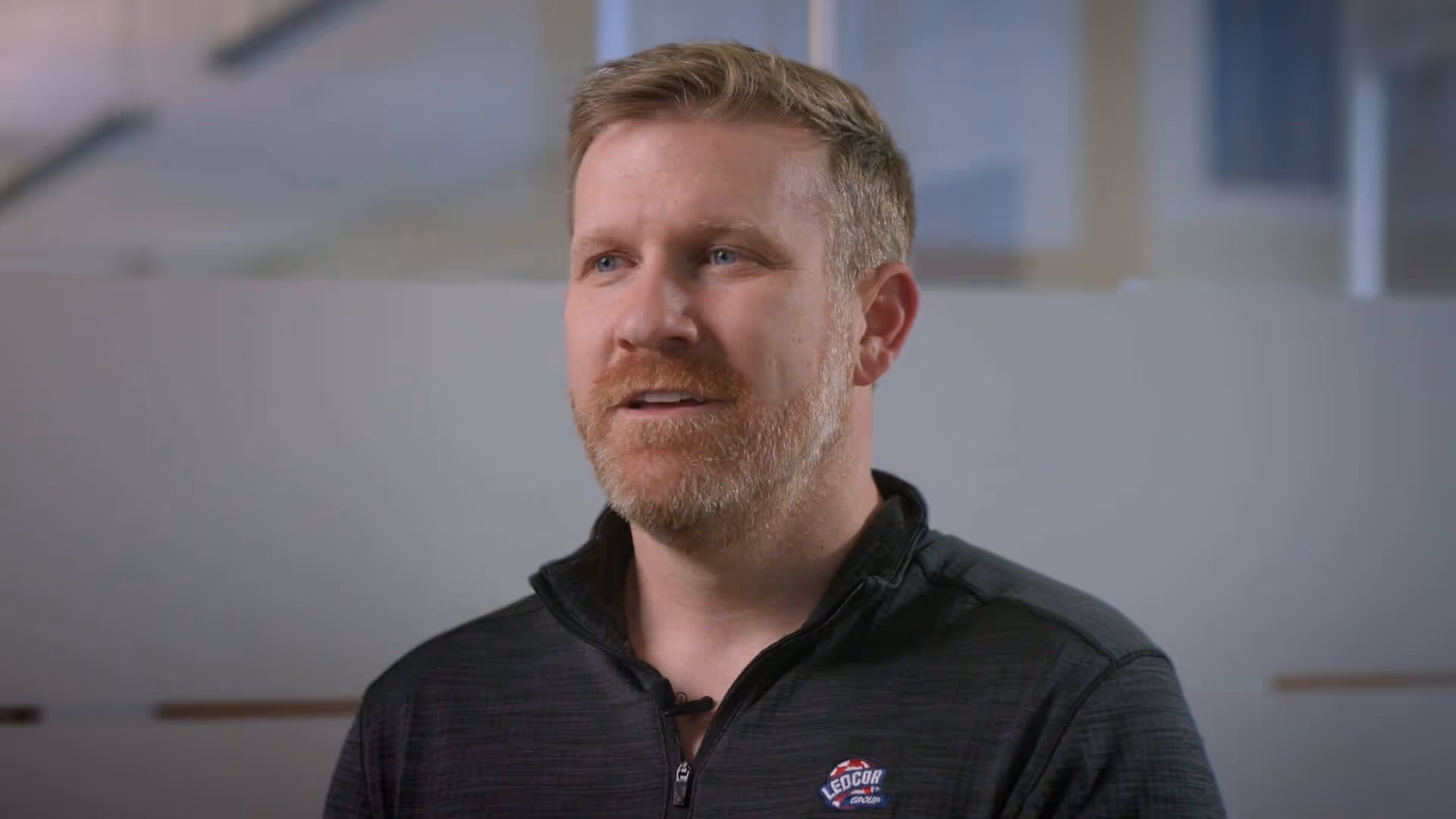 Man with blonde hair and beard wearing a black zip-up jacket with a LEGOCR Group logo, speaking indoors against a blurred background.