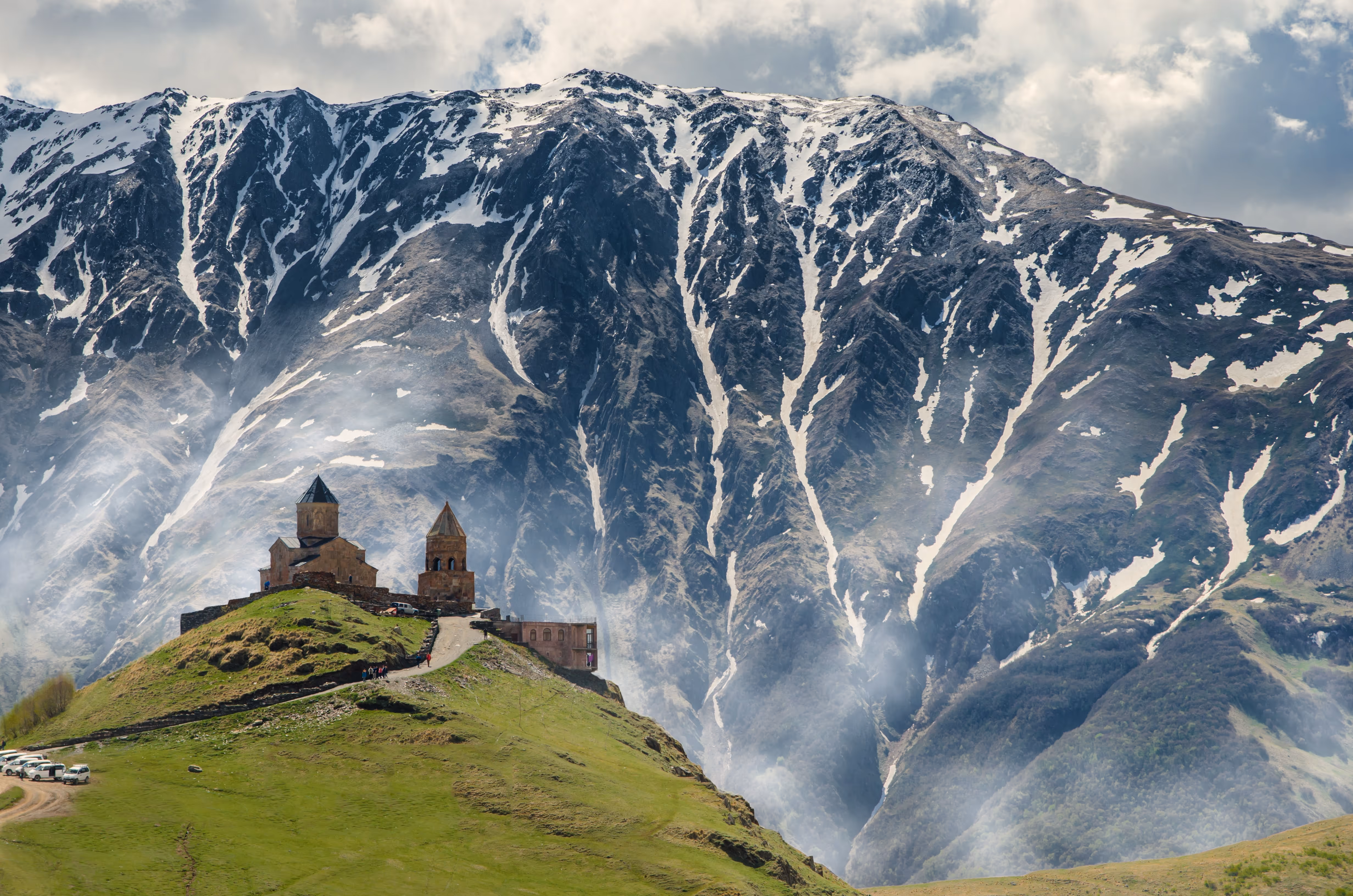 View of mountains covered in snow with clouds in the background and a house on a grassy hill in the foreground