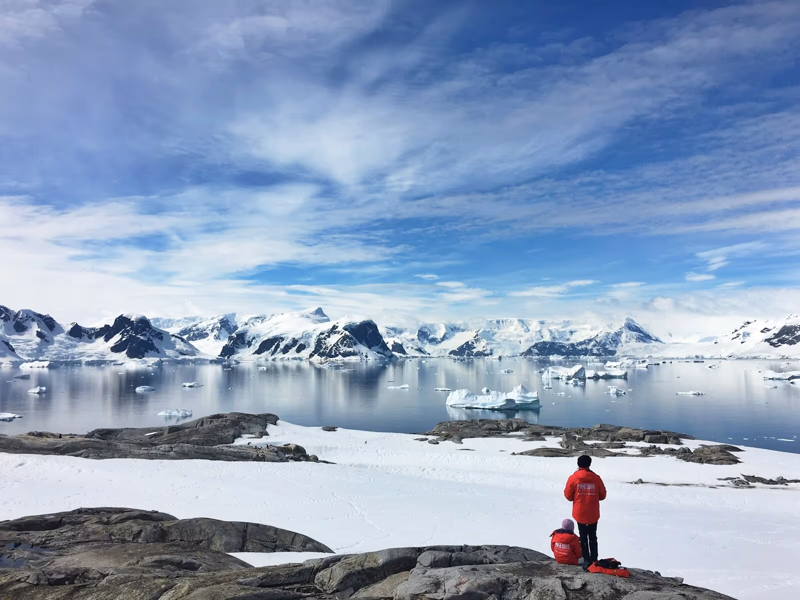 Two people in red jackets sitting on the ice looking over the icy landscapes of Antarctica
