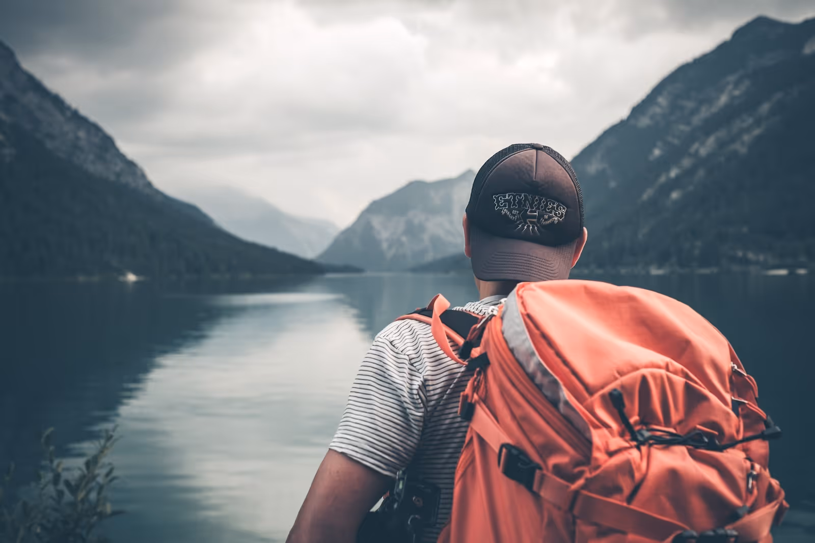 Person with a red backpack facing a lake surrounded by mountains