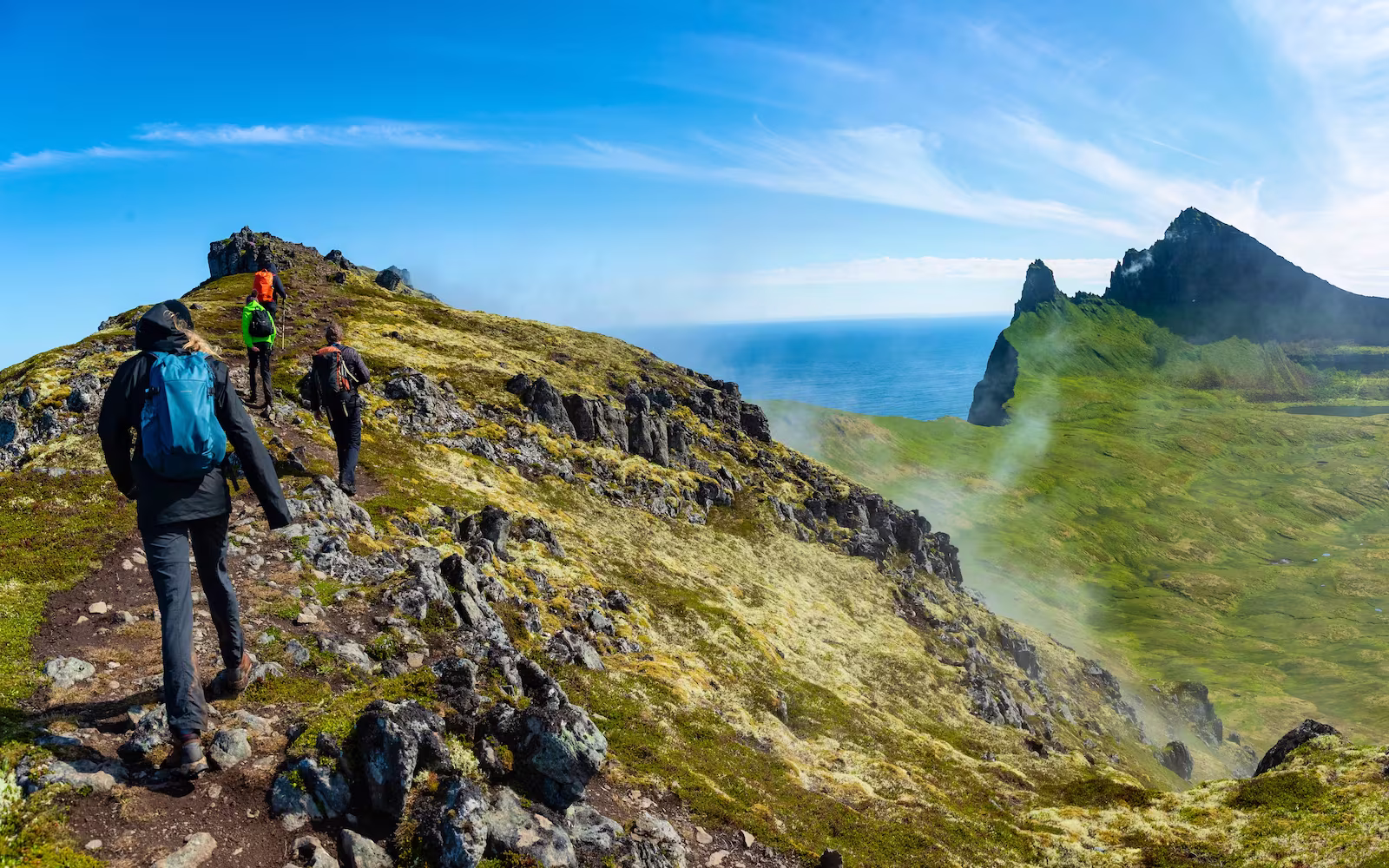 Hikers climbing green hills around volcano with ocean in background