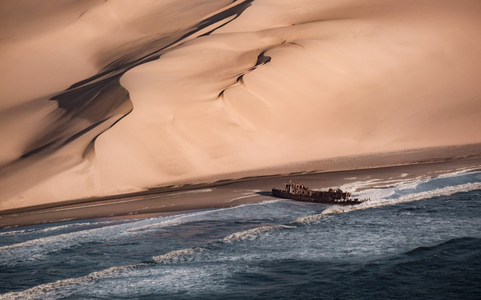 Shipwrecked boat in desert with ocean and sand mountains