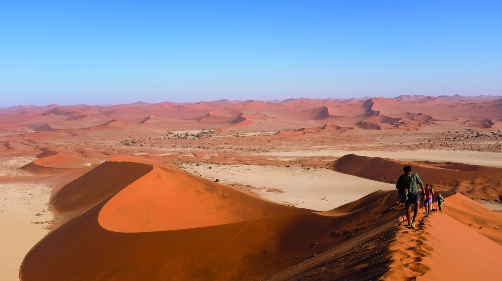 People climbing up sand dunes with blue skies and desert in background