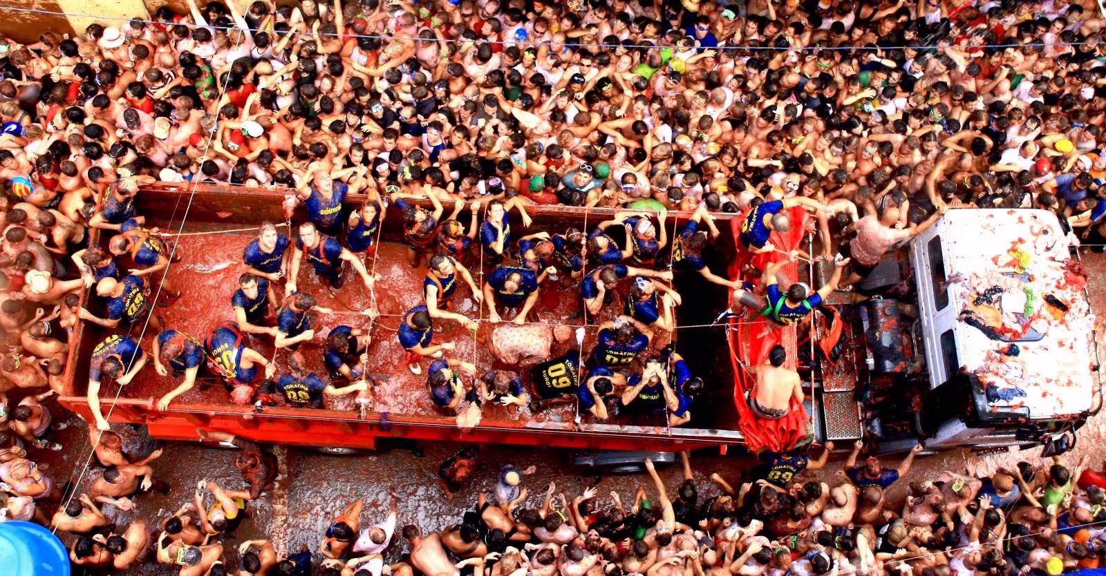 People in back of lorry with tomatoes, large crowd of people around lorry